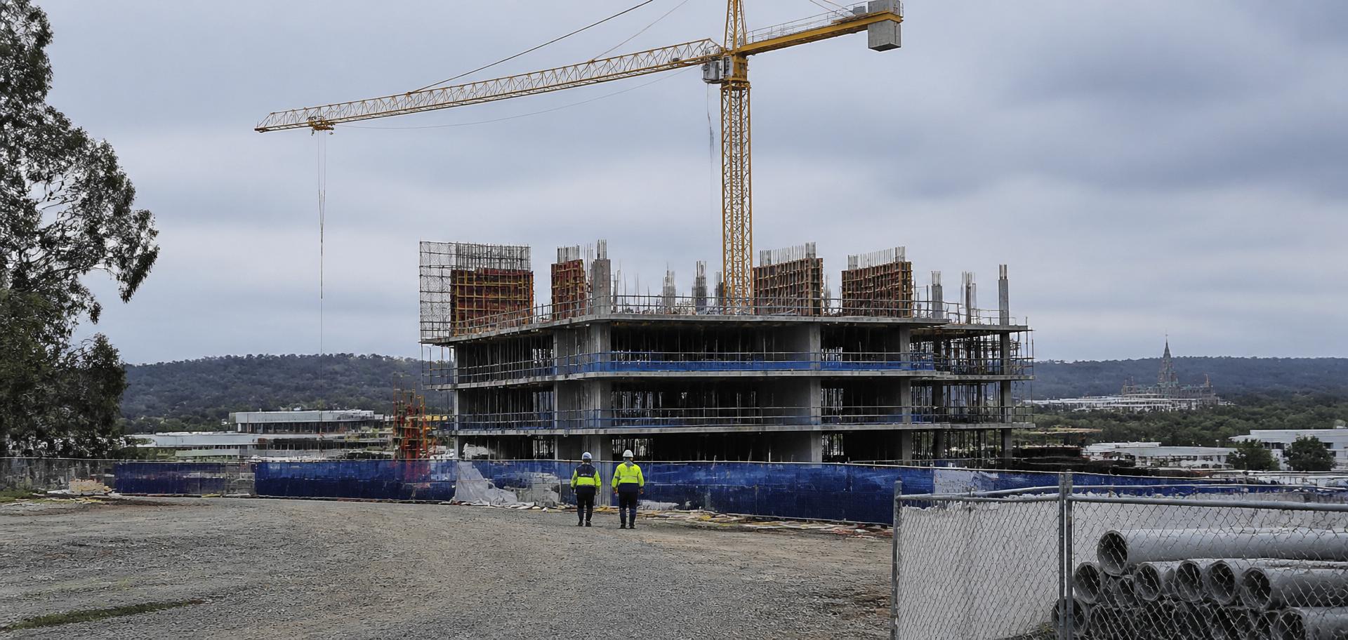 Mid-rise concrete-frame office building under construction with a yellow tower crane in a Canberra precinct, hi-vis workers walking the site, eucalypts and low bushland horizon behind — Spire Safety WHS consultants Canberra