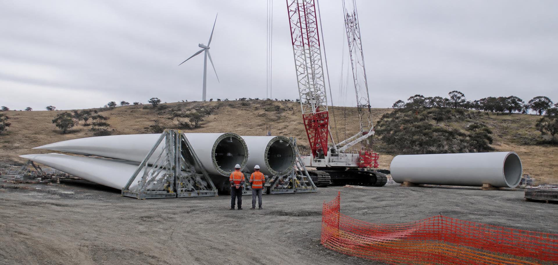 Wind farm construction laydown yard on the New England tablelands near Tamworth, NSW — three white turbine blades on steel transport cradles, a tower section on timber blocks, a red-and-white crawler crane rigging up and an erected turbine on the ridge behind, with two workers in orange hi-vis and hard hats standing at the blade root and dry golden tablelands paddocks beyond — Spire Safety WHS consultants Tamworth