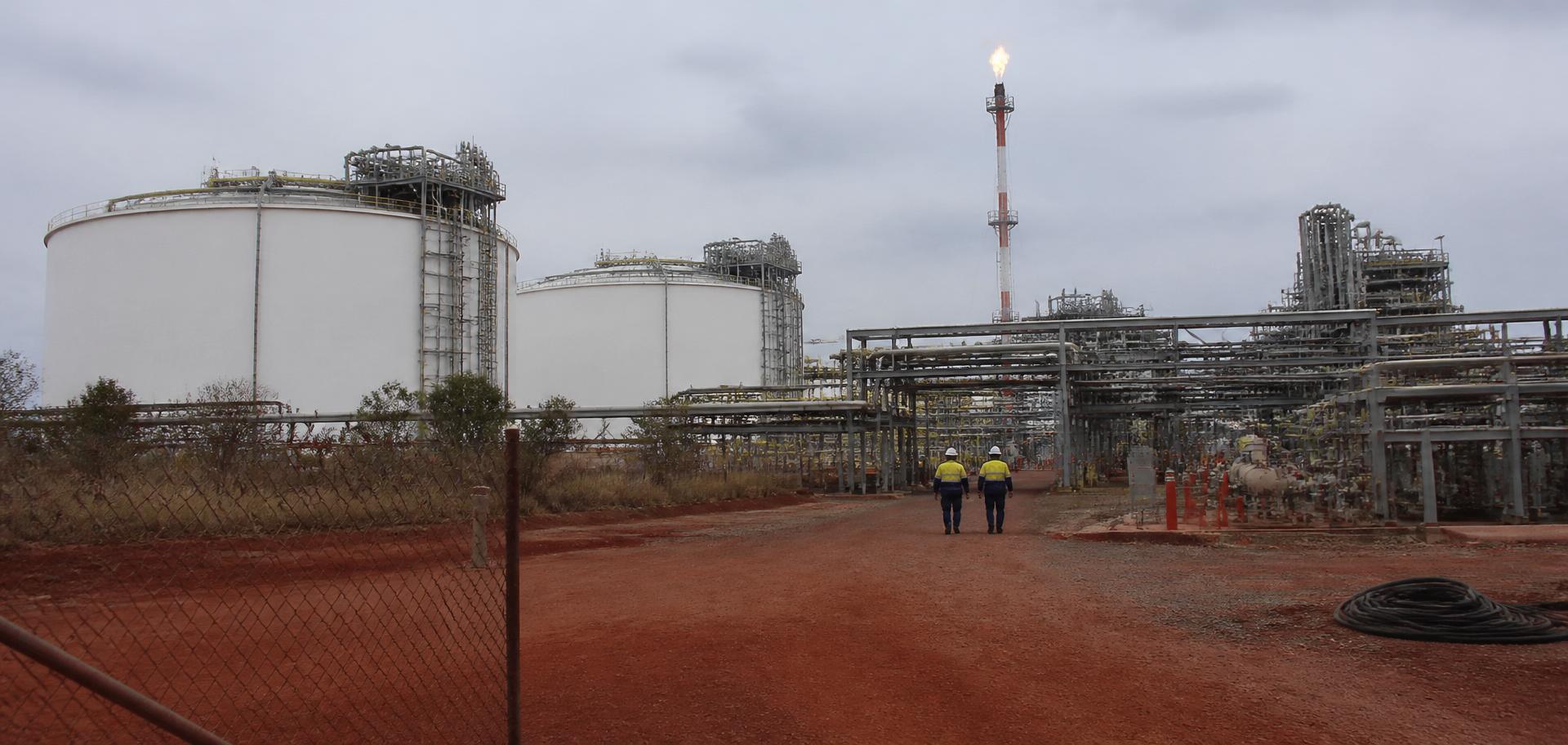 Onshore LNG processing plant outside Darwin — white cryogenic storage tanks, silver pipe-racks and a flare stack with two hi-vis workers walking through on red laterite soil — Spire Safety WHS consultants Darwin
