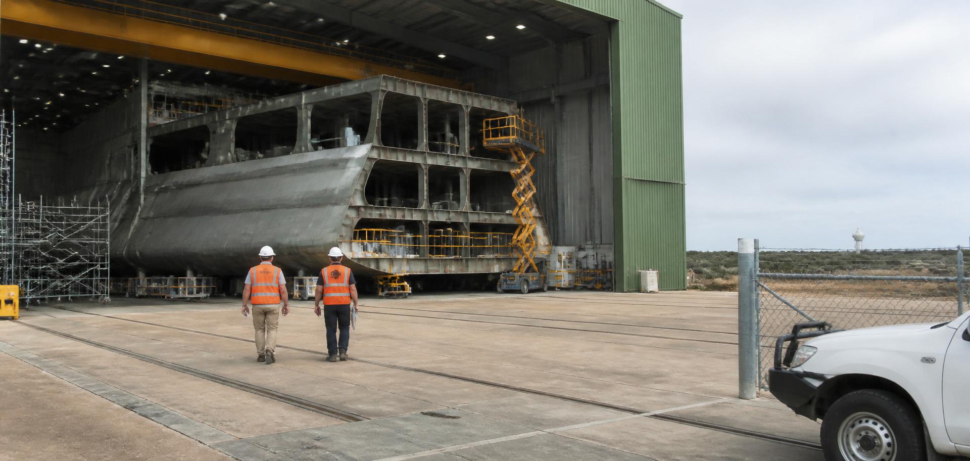 Grey steel naval hull section under construction inside a green-roofed shipbuilding hall at the Osborne Naval Shipyard, Adelaide — Spire Safety WHS consultants Adelaide
