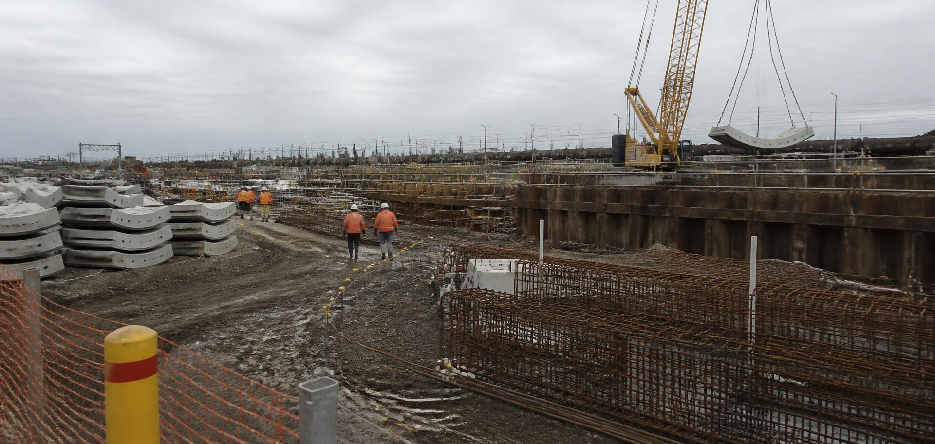 West Gate Tunnel Project construction site in Melbourne — stacked precast concrete tunnel segments, a large yellow crawler crane lifting a segment, workers in orange hi-vis on a muddy site with rebar cages and freight rail lines on the horizon — Spire Safety WHS consultants Melbourne