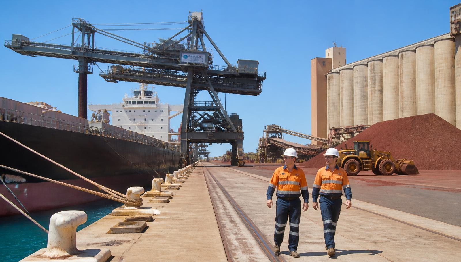 Bulk export wharf at the Port of Geraldton, Mid West Western Australia — a steel ship loader with conveyor gantry over a moored bulk carrier, tall concrete grain silos and a rust-red iron ore stockpile with a front-end loader working the yard, and two workers in orange hi-vis and hard hats walking the concrete apron beside the mooring bollards, blue Indian Ocean water alongside — Spire Safety WHS consultants Geraldton