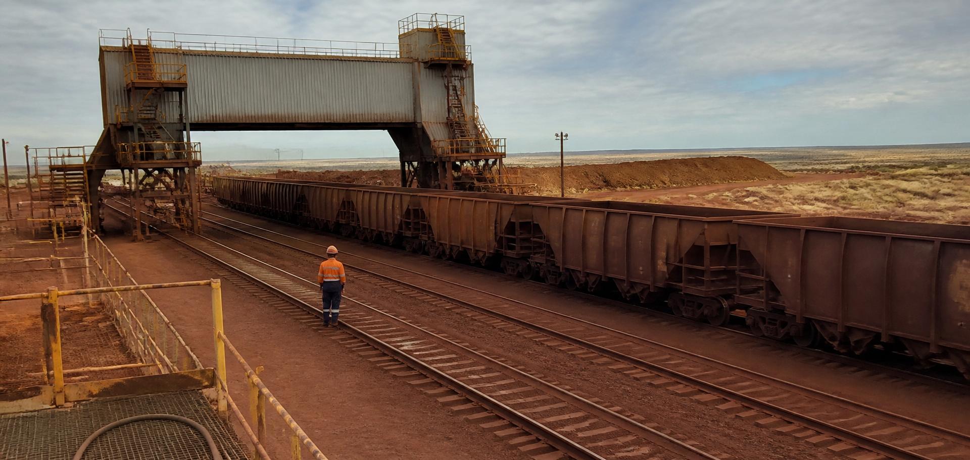 Iron-ore rail loader and dark rust-red ore wagons on red Pilbara earth, Western Australia — Spire Safety WHS consultants Perth